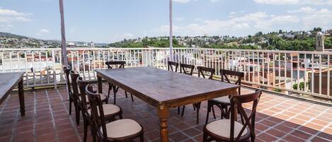 Una terraza con mesa y sillas de madera, baranda blanca y vista panorámica de la ciudad.