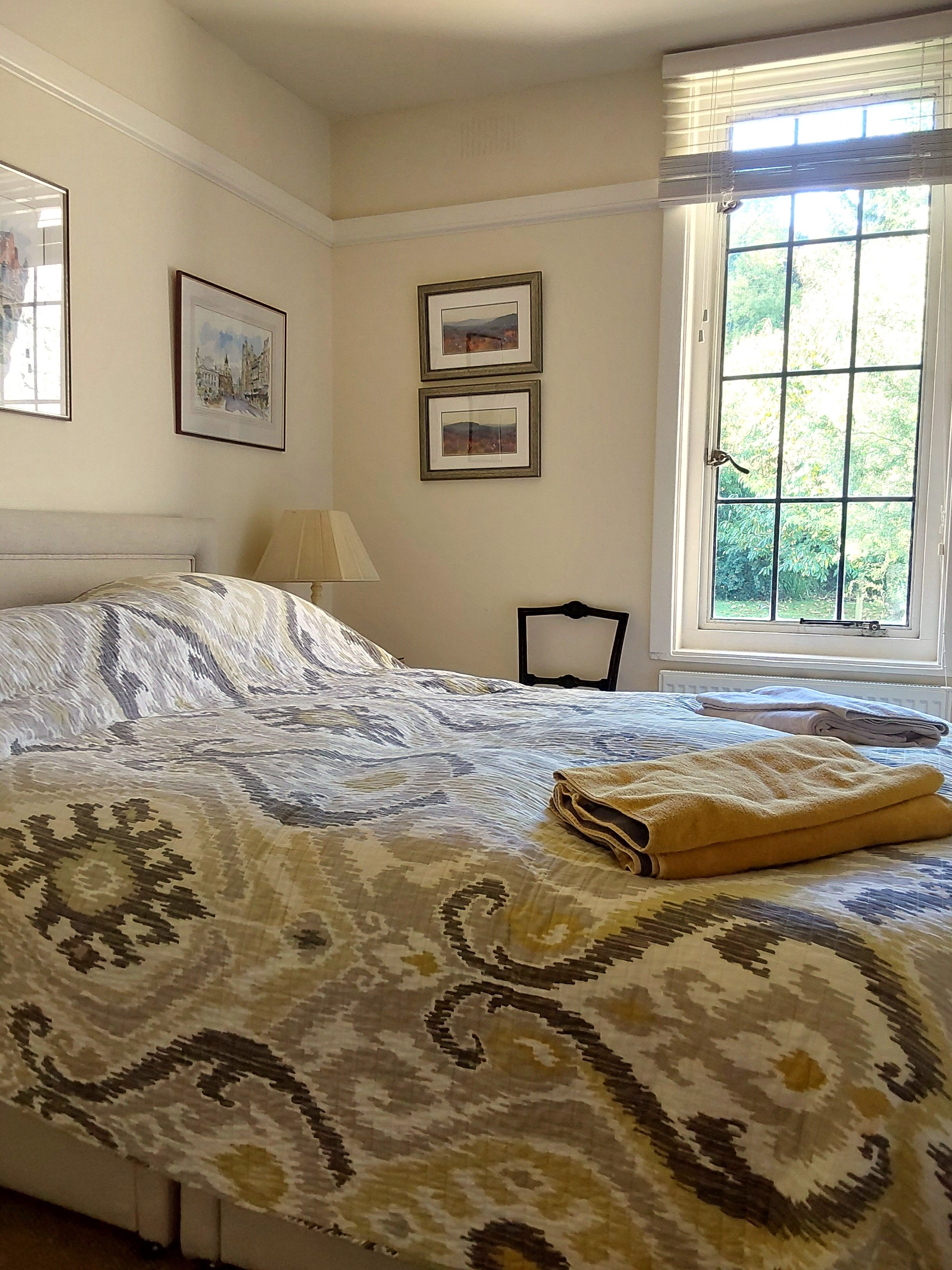 A bedroom with a bed, two framed pictures on the wall, a window with blinds, and a lamp.