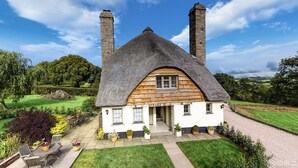 A thatched-roof house with a stone chimney, surrounded by a well-maintained garden and a paved driveway. - Rock House Cottage (Exeter)
