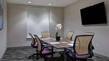A conference room with a long table, six chairs, a potted plant, a whiteboard, and a flat-screen TV.
