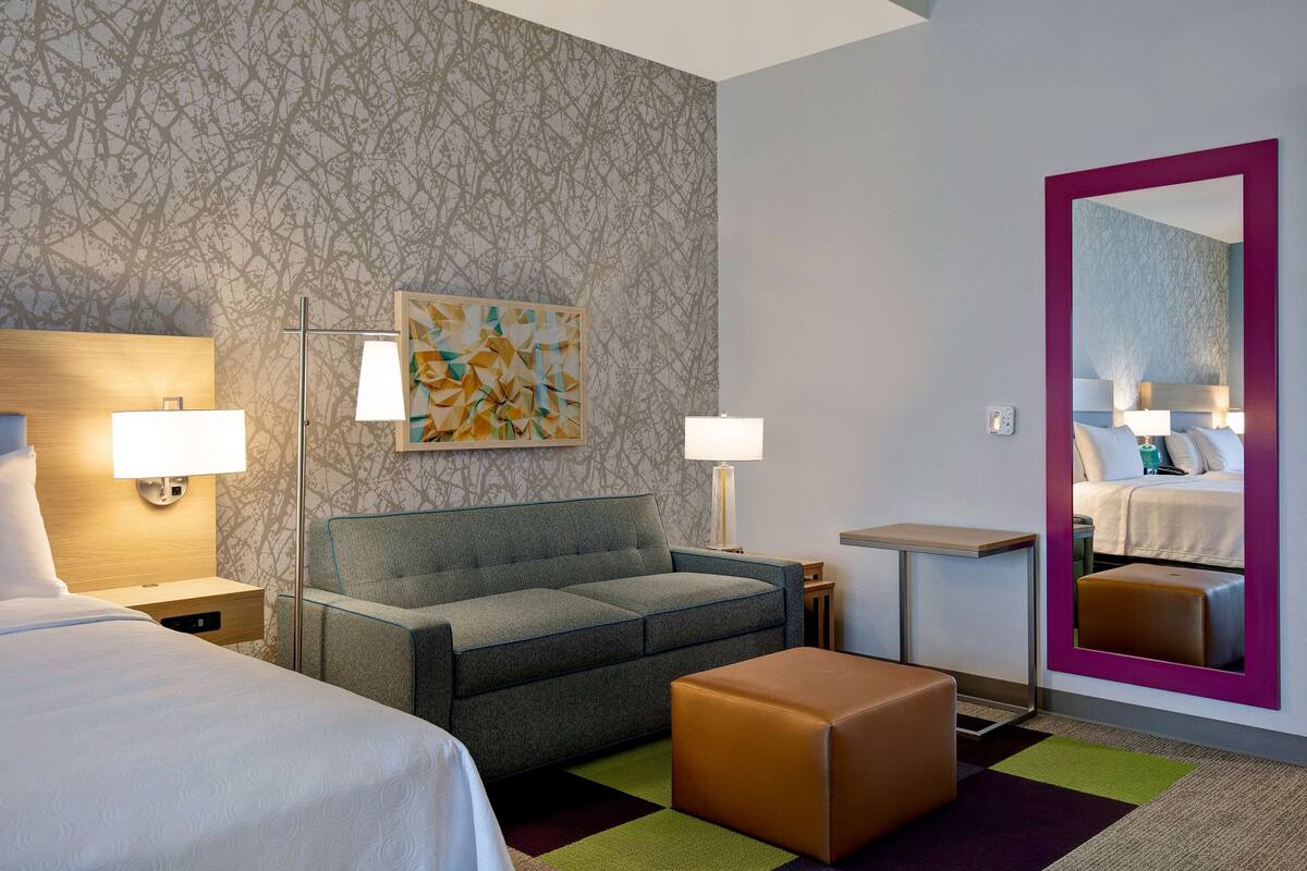 A hotel room with a bed, a sofa, a small table, a chair, a mirror, and a painting on the wall.