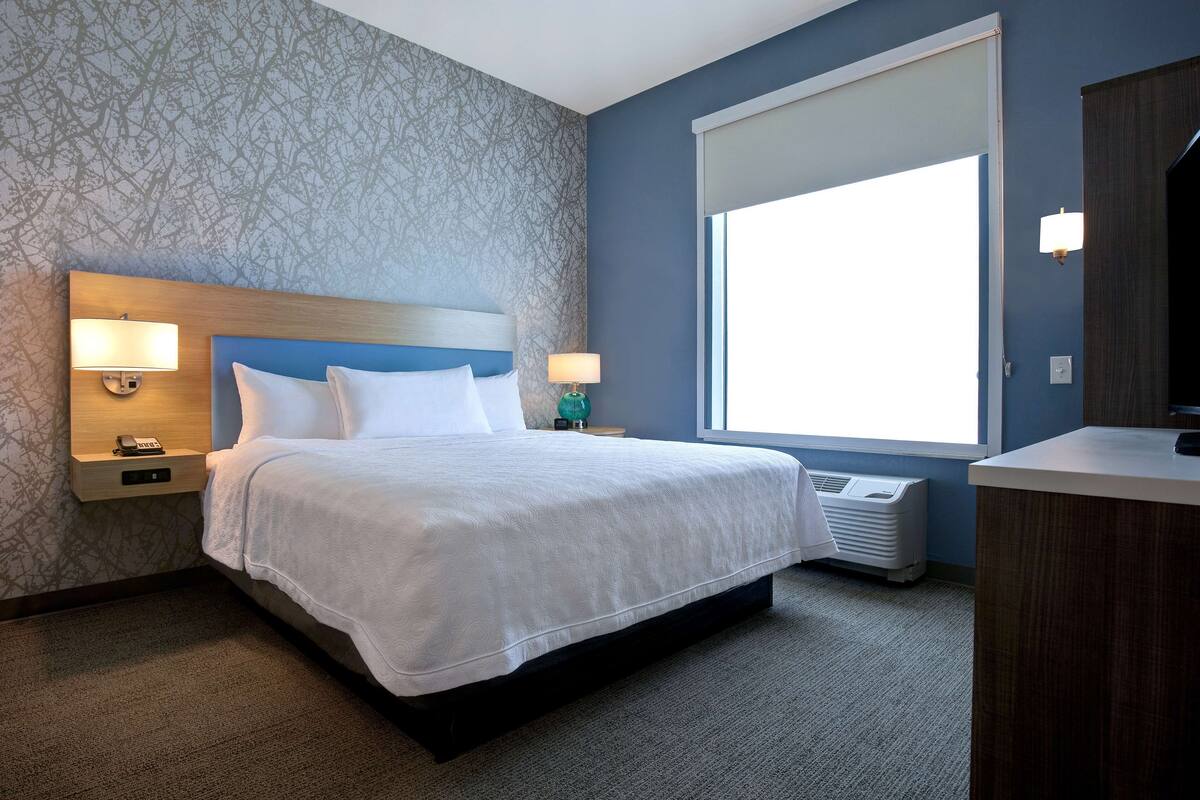 A hotel room with a large bed, two bedside lamps, a nightstand, a window, and a built-in desk with a lamp.