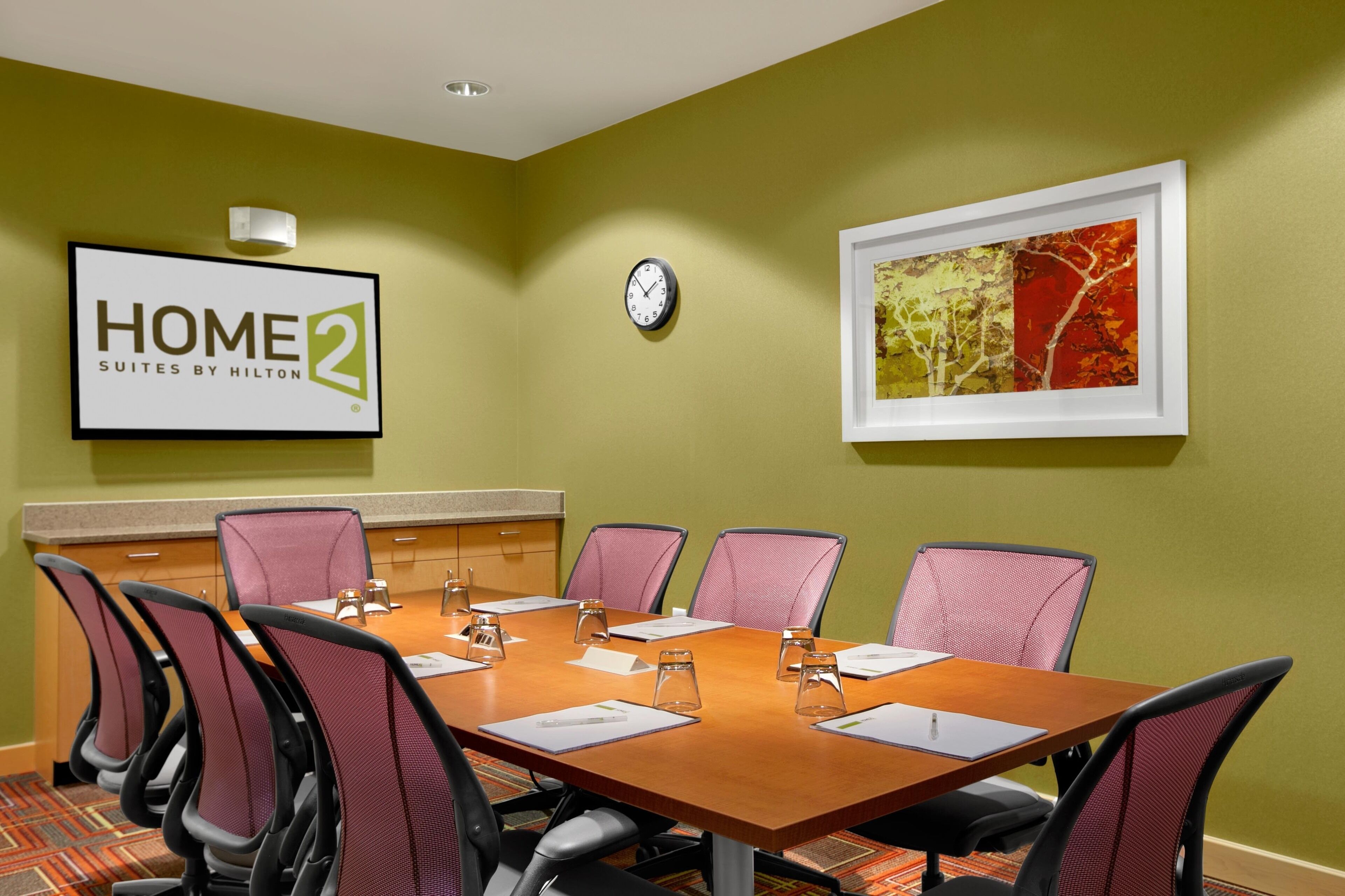 A conference room with a long table, six chairs, a clock, and a framed picture on the wall.
