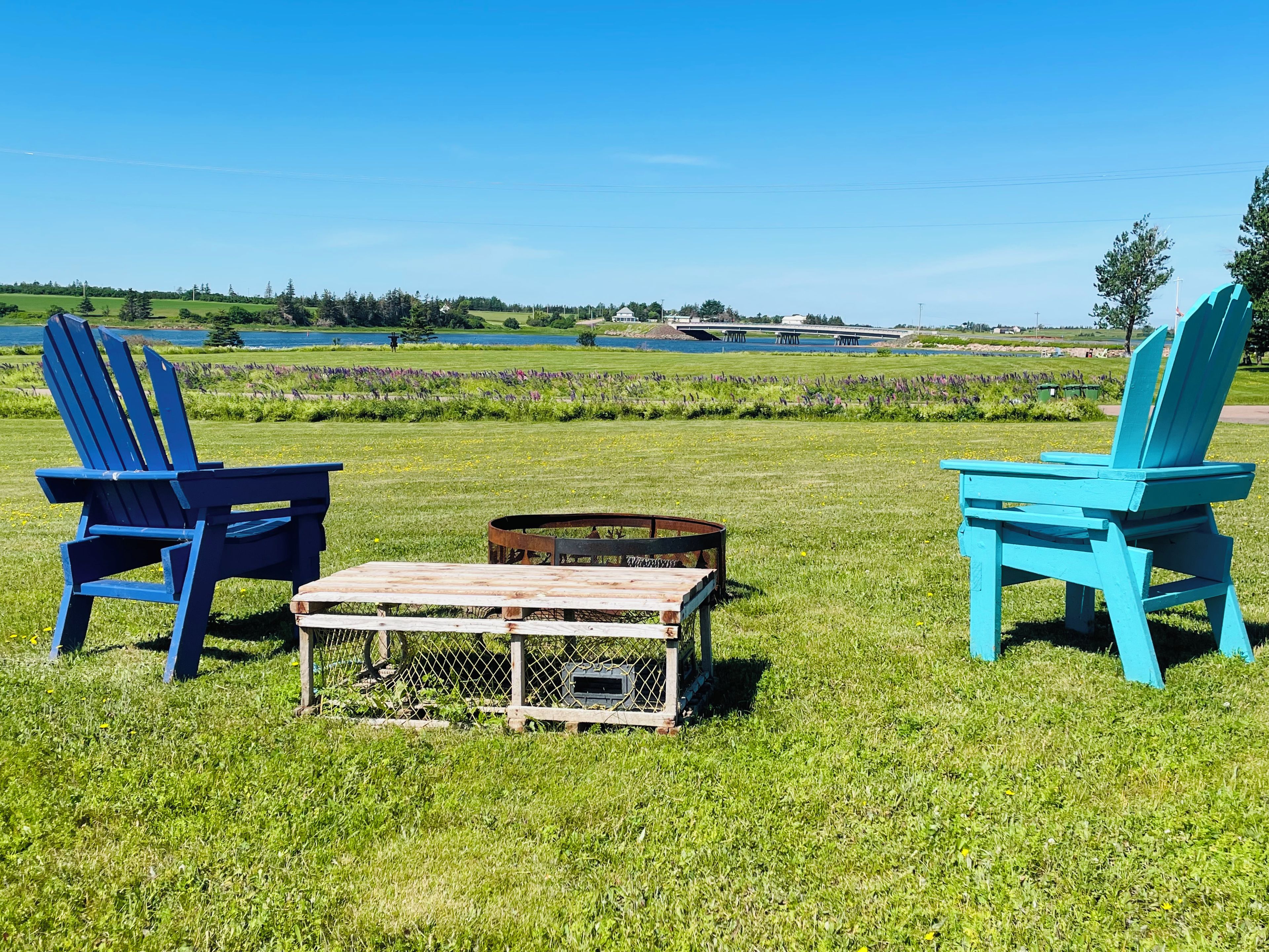 Deux chaises Adirondack bleues, une table en bois et un foyer métallique, le tout installé dans un champ verdoyant avec un lac et un pont en arrière-plan.