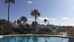A clear blue pool with a metal railing, surrounded by palm trees and buildings in the background.