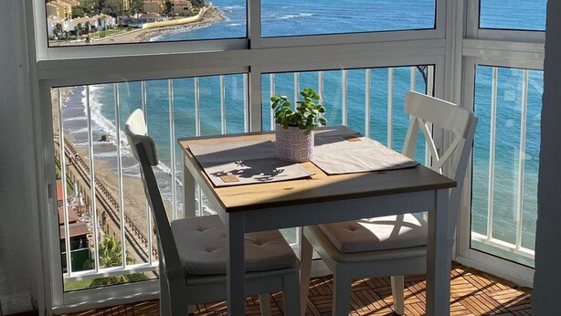 A balcony with a table and chairs, overlooking a coastal view with buildings and the sea.
