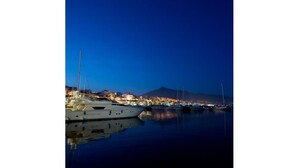 A marina at night with boats docked, calm water, and a cityscape with buildings and a mountain in the background.