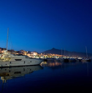 A marina at night with boats docked, calm water, and a cityscape with buildings and a mountain in the background. - Admiral Building Studio (Mijas)