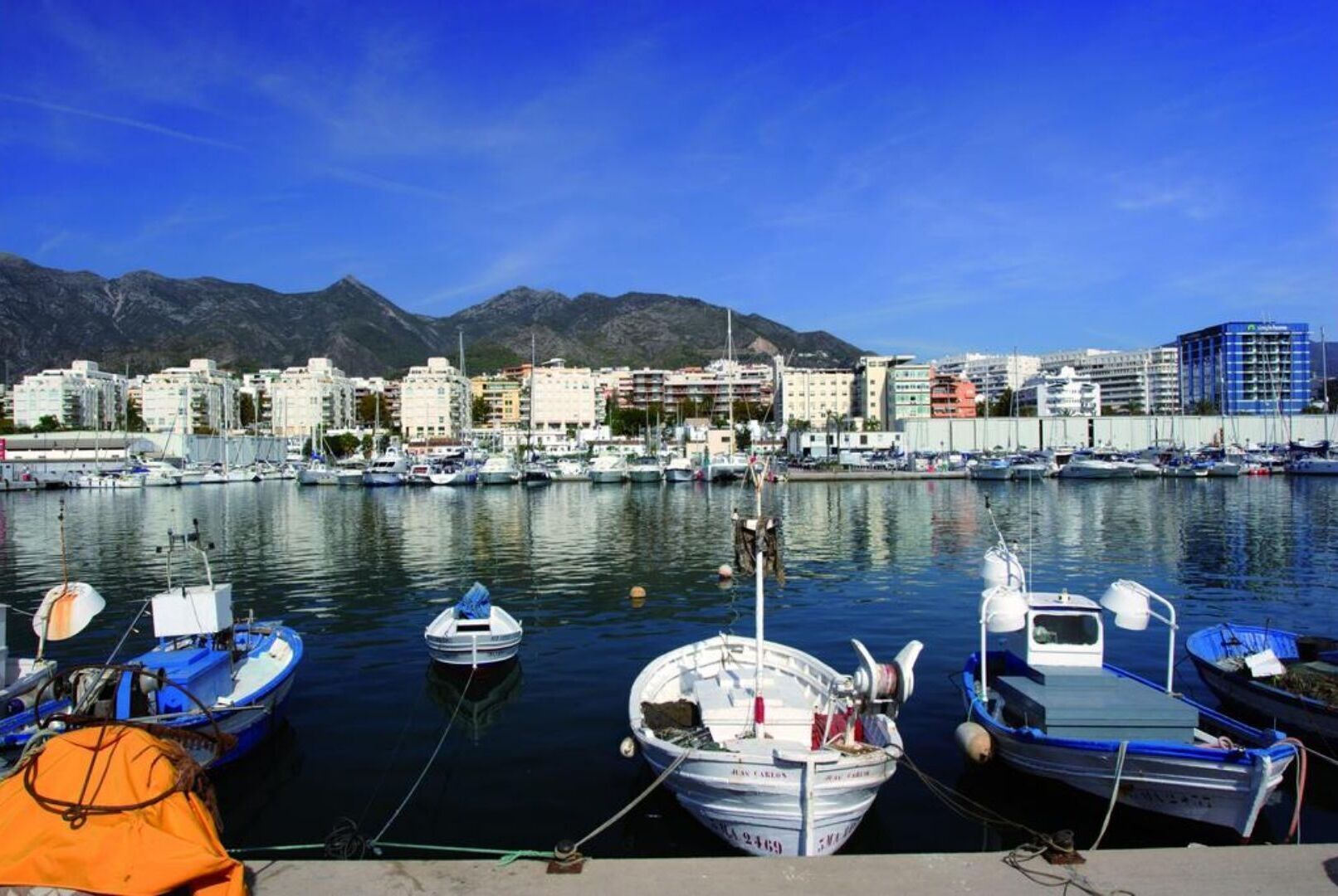 Un port de plaisance avec des bateaux amarrés, un paysage urbain avec des bâtiments en arrière-plan et des montagnes à l’horizon.