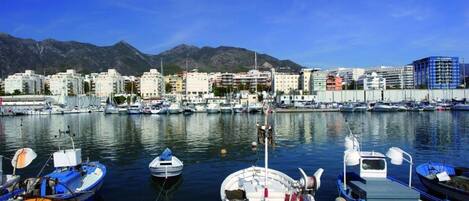 A marina with boats docked, a cityscape with buildings in the background, and mountains in the distance.