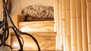 Una escalera de madera que lleva a una cama, cortinas de bambú y una pared con un ventanal circular.