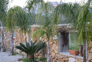 Un edificio rústico de piedra con techo de paja, rodeado de palmeras y plantas en macetas.