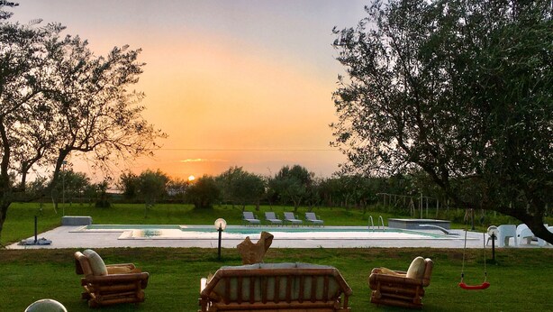 Zona de piscina con sillones de descanso y un banco de madera, rodeada de árboles y vistas al atardecer.