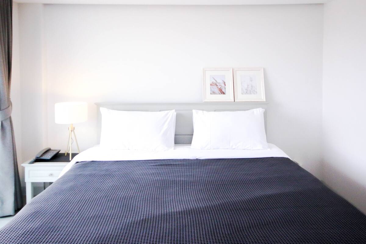 Ein Hotelzimmer mit einem ordentlich bezogenen Bett, einem Nachttisch mit Lampe und zwei gerahmten Bildern an der Wand.
