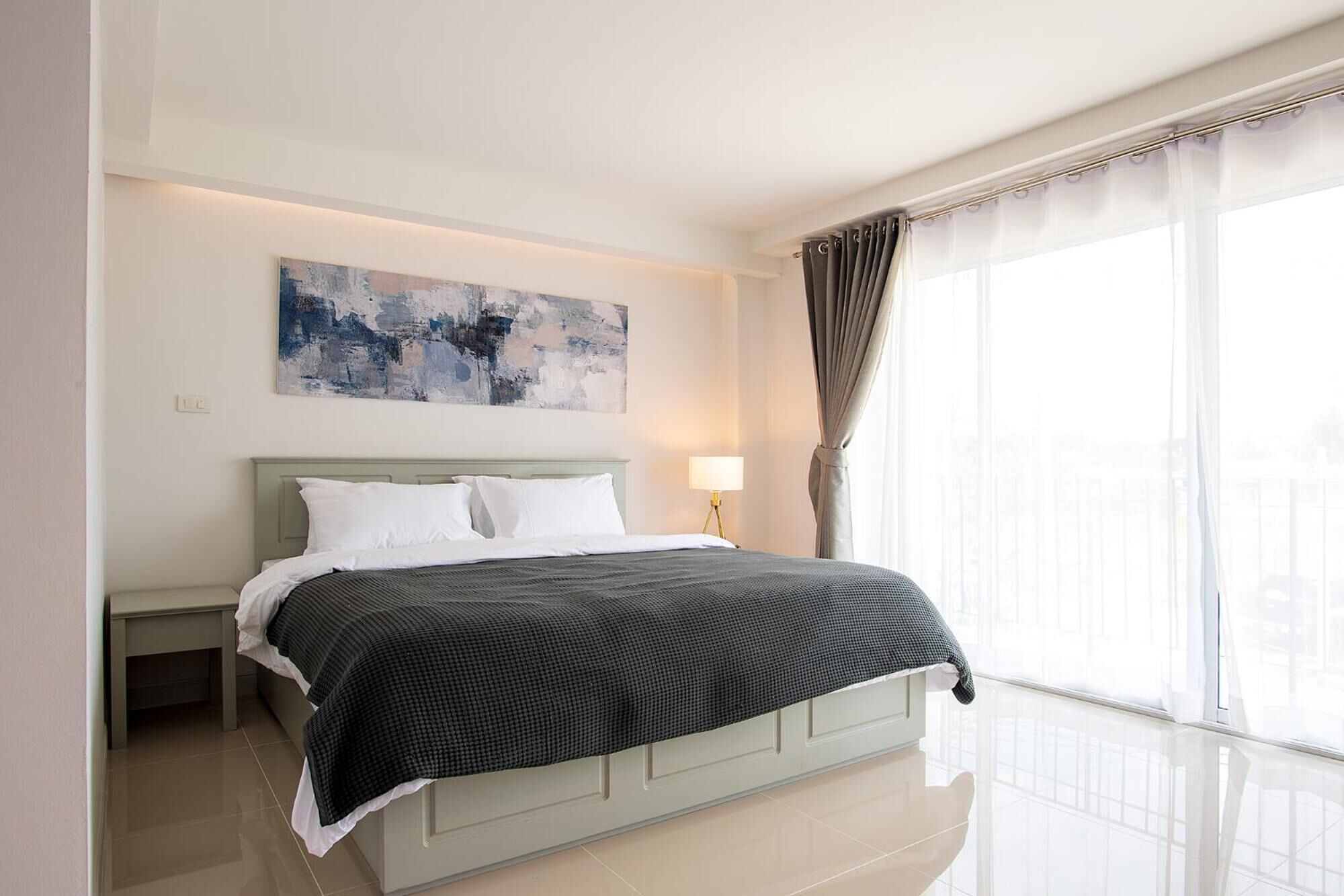 Un hotel moderno con una cama grande, una lámpara de noche, un cuadro en la pared y vistas de la ciudad a través de cortinas translúcidas.