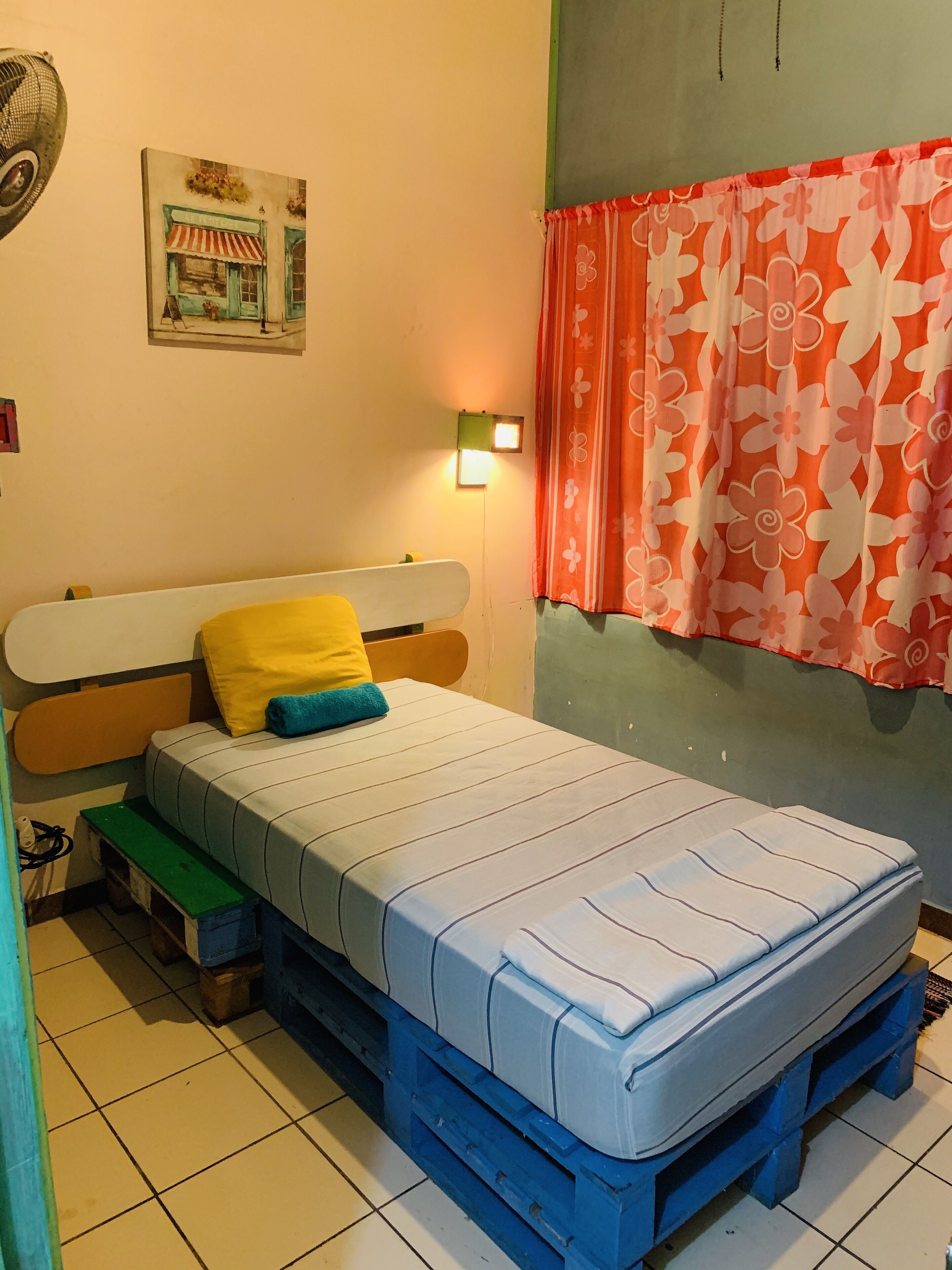 A room with a bed on a blue pallet, a wall-mounted light, a floral curtain, and a painting of a street scene.