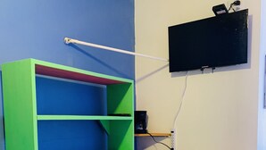 Una habitación con una estantería verde y azul, un escritorio blanco con cajones azules, un televisor de pantalla plana negro montado en la pared y una cafetera sobre una repisa.