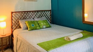 Una cama bien hecha con una manta verde y almohadas, un cabecero de madera, un aire acondicionado montado en la pared y un cuadro con una escena callejera.