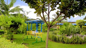 A playground with a swing set and climbing structures, surrounded by lush greenery and cacti.
