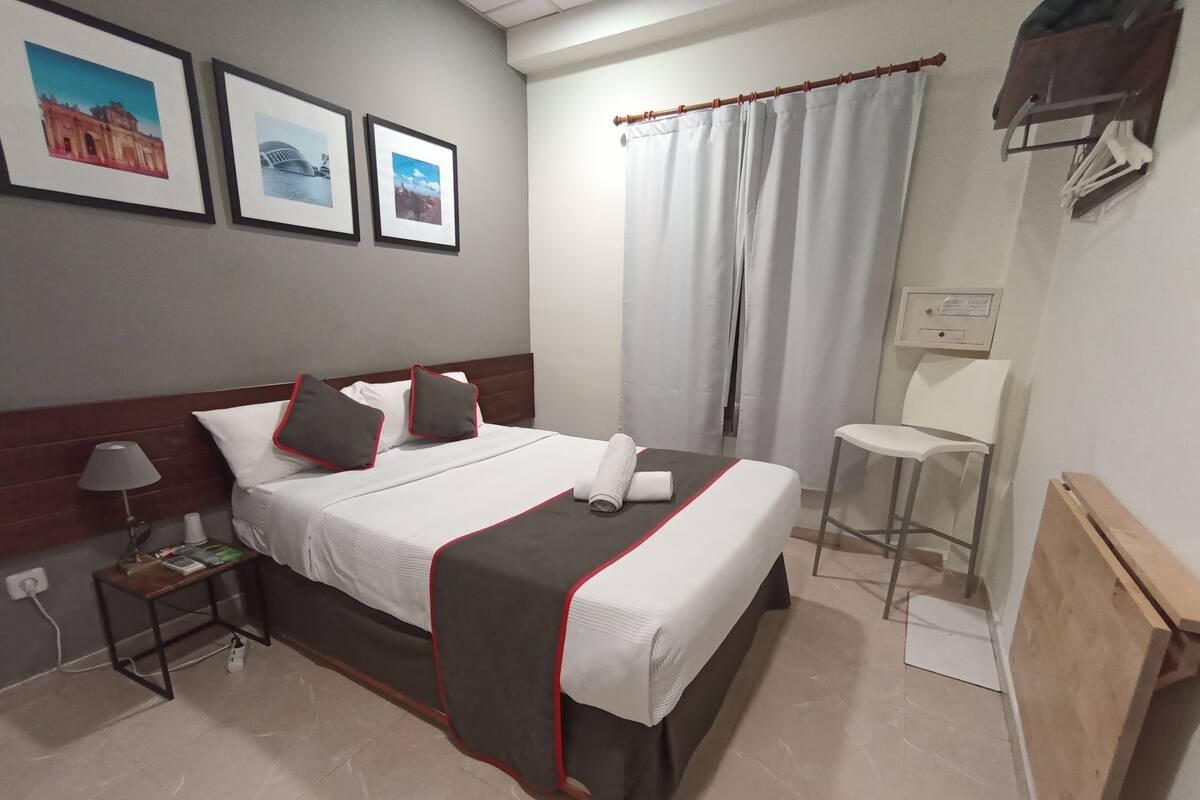 Una habitación de hotel con cama, mesita de noche, silla, cortina e imágenes enmarcadas en la pared.