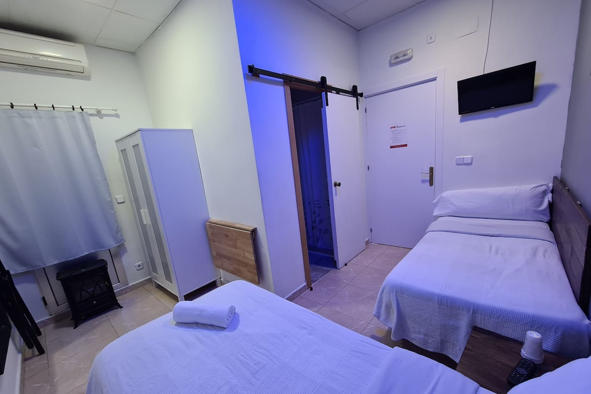 Habitación de hotel con dos camas individuales, un televisor montado en la pared, una puerta de madera y un pasillo iluminado en azul.