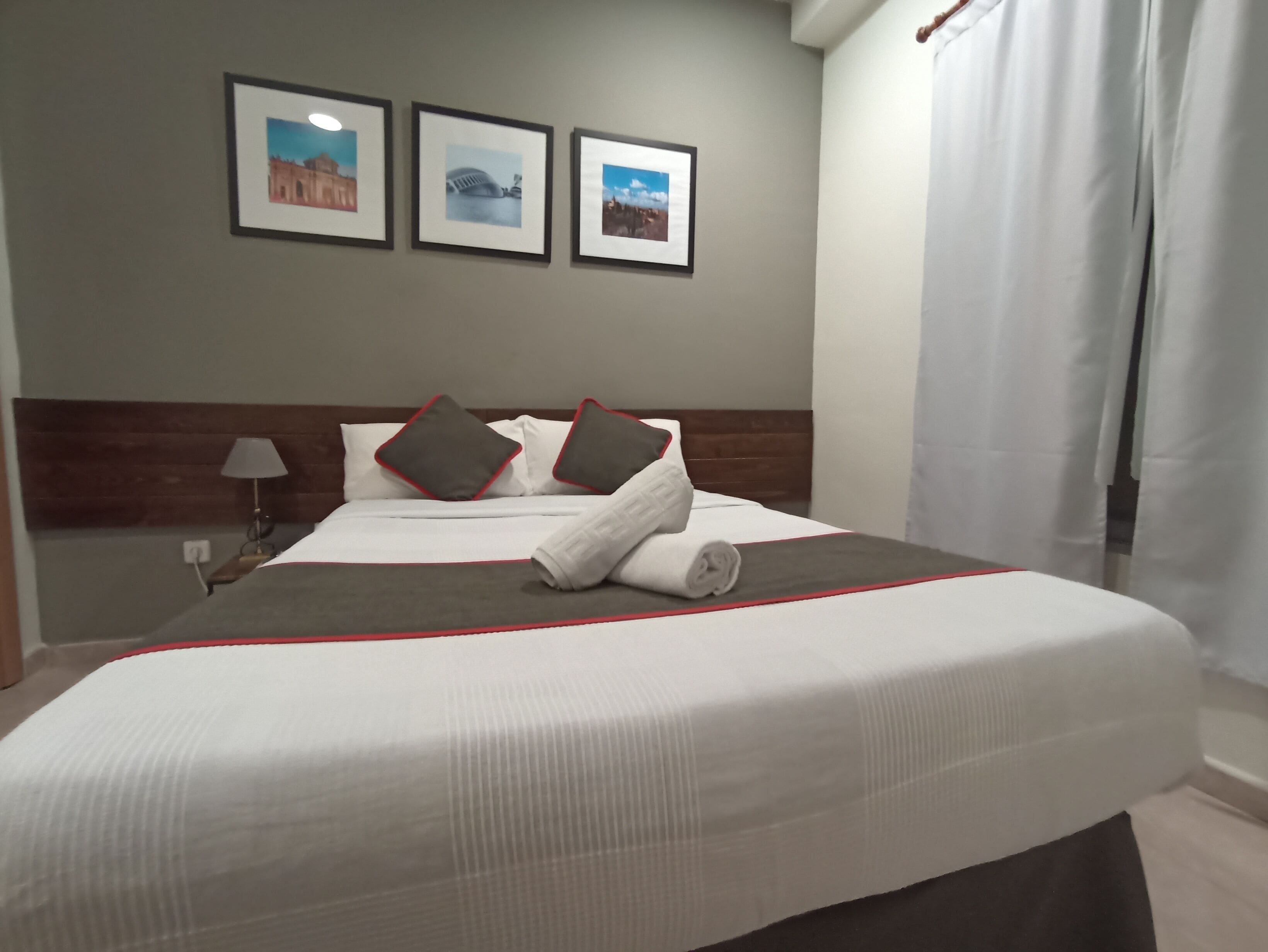 A neatly made bed with a white and gray bedspread, two pillows with red trim, and a rolled towel on the bed. There are three framed pictures on the wall above the bed.