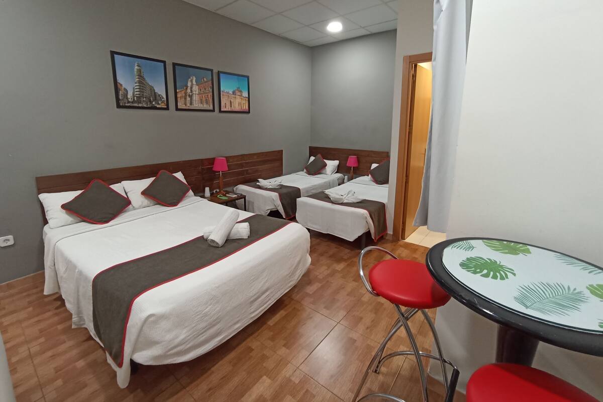 Habitación de hotel con dos camas, una mesita y una silla. Hay cuadros en la pared y una puerta que da a otra habitación.