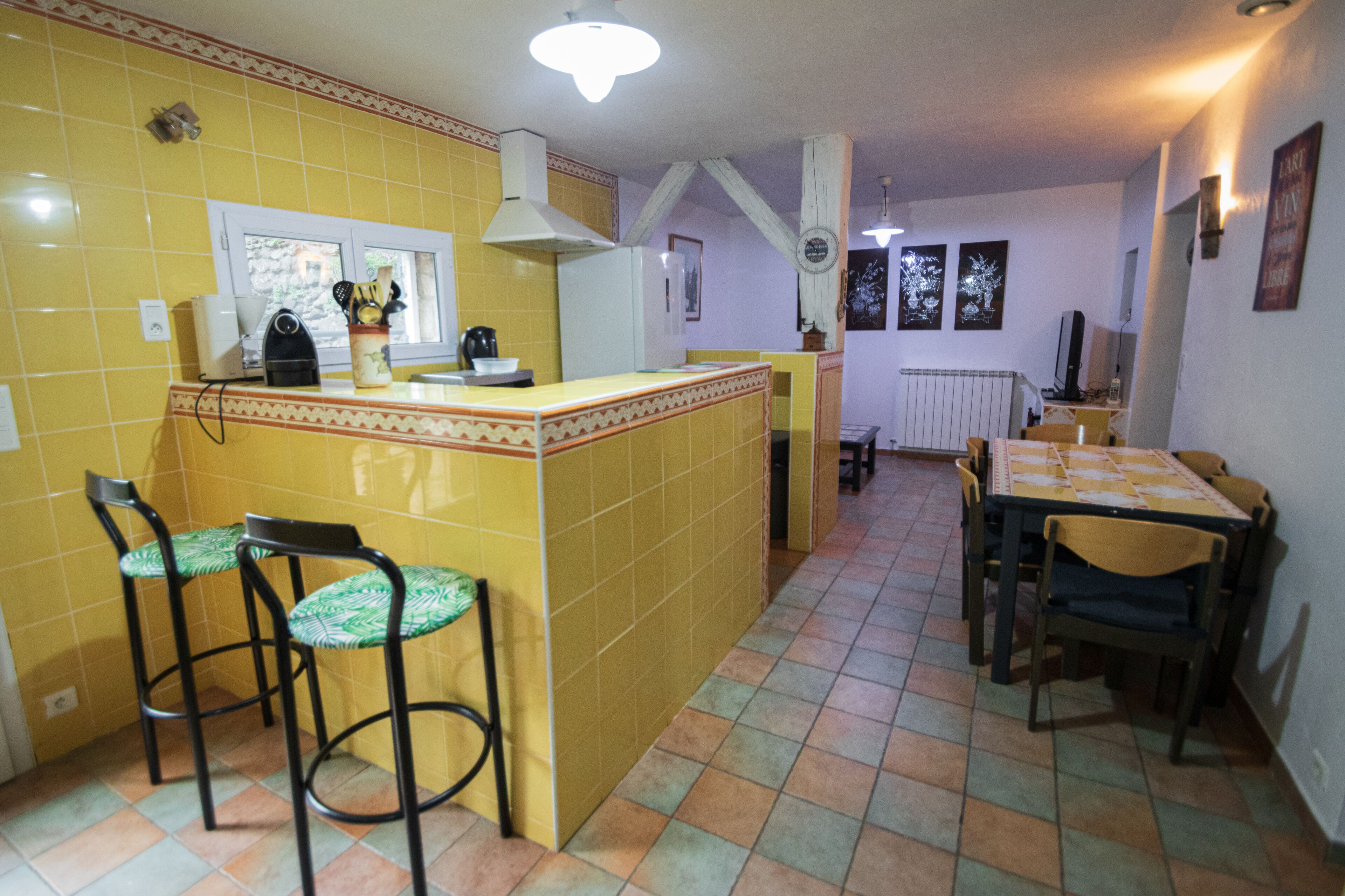 Une cuisine avec des carreaux jaunes, un îlot central doté de tabourets, une table à manger avec des chaises et une fenêtre offrant une vue agréable.