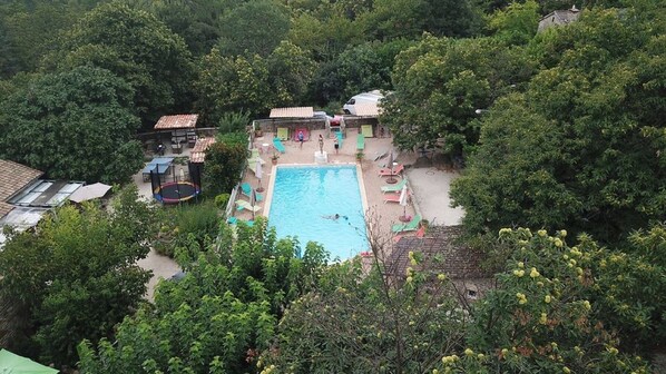 An aerial view of a resort with a swimming pool surrounded by greenery, a trampoline, and several small buildings.