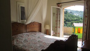 Ein Schlafzimmer mit Bett, Nachttisch, Lampe und Blick auf Grünflächen und Gebäude durch eine offene Tür.