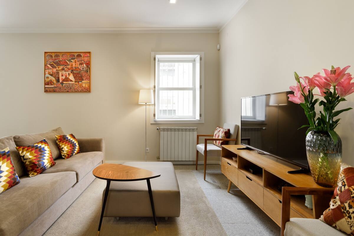 A living room with a sofa, ottoman, TV, and a vase with flowers.