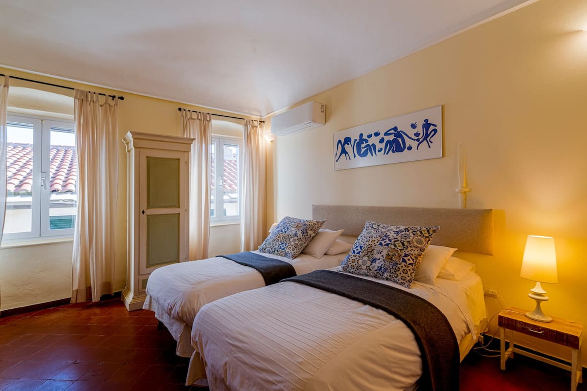 Habitación de hotel con dos camas, una mesita de noche con lámpara, un cuadro en la pared y una ventana con cortinas.