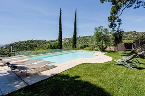 Una piscina con sillones de descanso, rodeada de un césped cuidado y altos árboles.