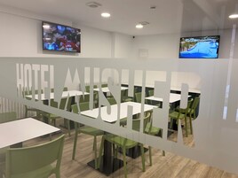 Un comedor de hotel con mesas y sillas, dos televisores montados en la pared y un amplio panel de vidrio esmerilado con la palabra 'HOTEL' parcialmente visible.