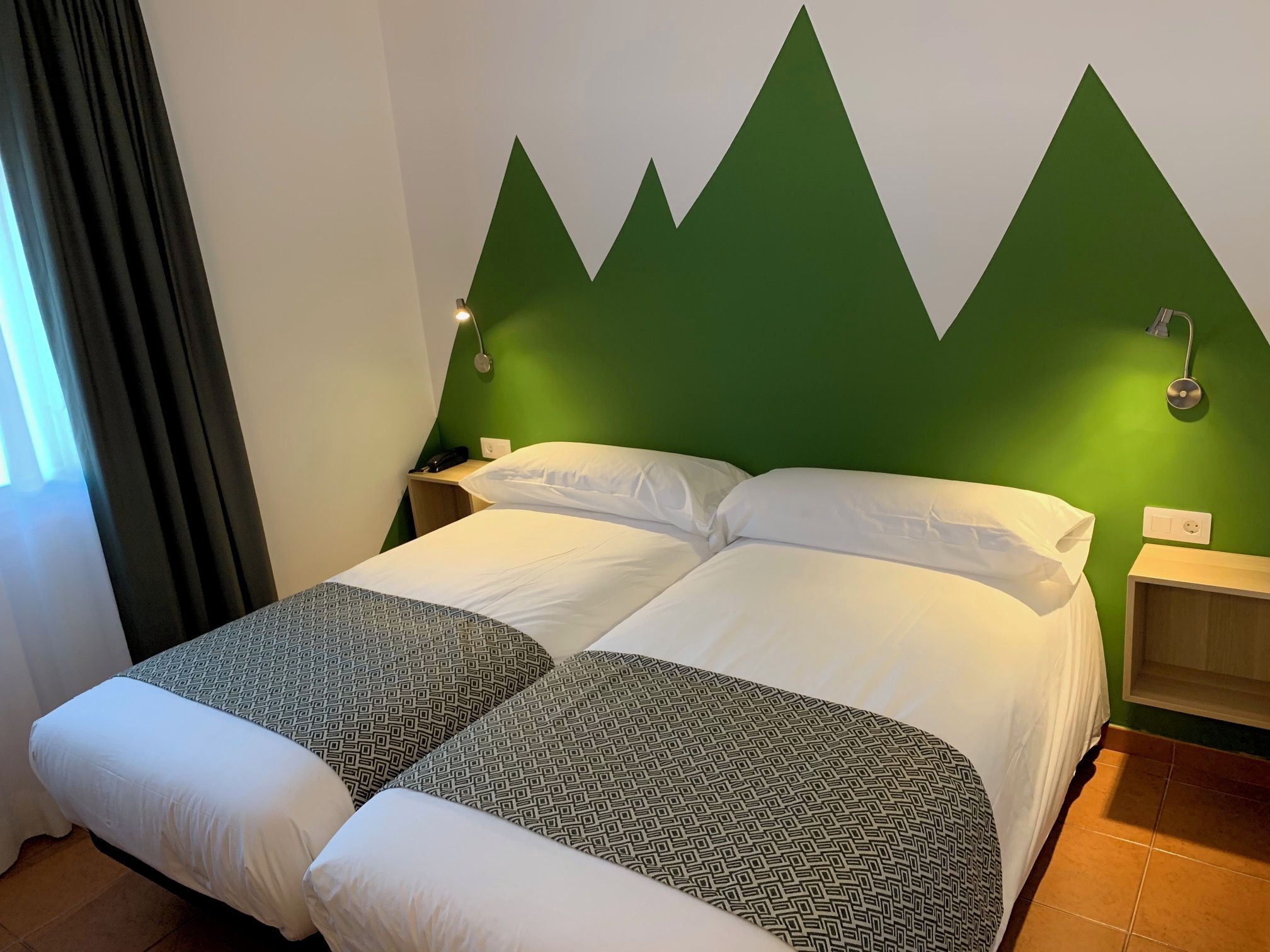 A double bed with a geometric patterned bedspread, a green accent wall, bedside lamps, and a nightstand with a phone.