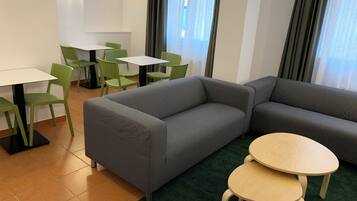 A modern hotel room with a grey sofa, two small round tables, green chairs, and a large window with curtains.
