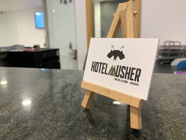 Une pancarte sur un chevalet en bois portant l’inscription « HÔTEL MUSHER » et « PAS DE LA CASA – ANDORRE », indiquant le nom et l’emplacement de l’hôtel.