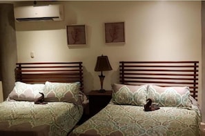 A bedroom with two beds, each with a patterned bedspread and a wooden headboard. There is a wall-mounted air conditioner above the beds and two framed pictures on the wall.