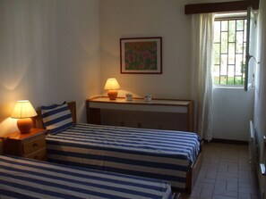 A hotel room with two beds, a desk, a lamp, a painting, and a window with curtains.