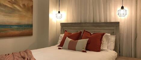 A neatly made bed with white and red pillows, a wooden headboard, and a painting of a sunset on the wall.