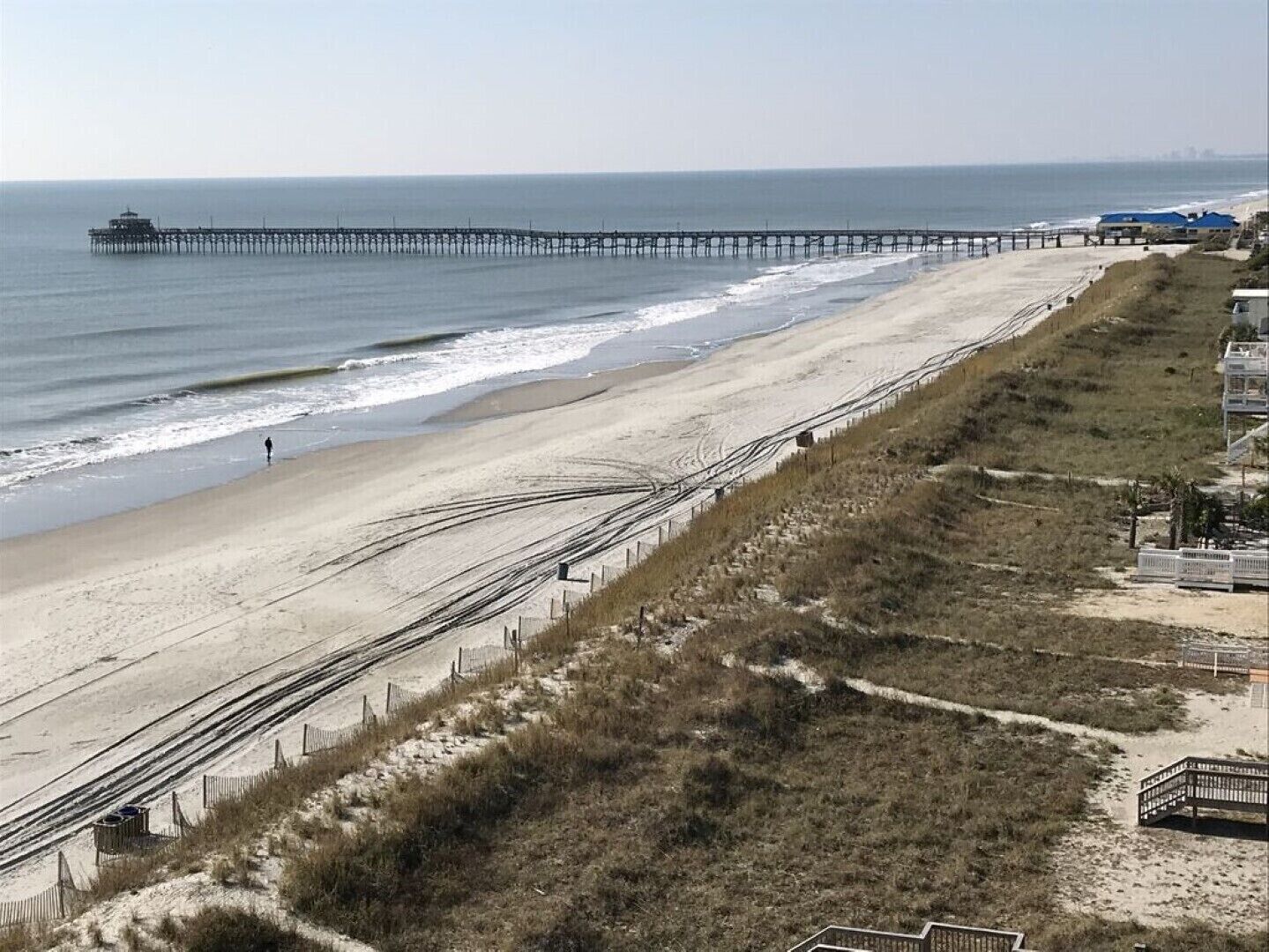 Une plage avec une longue jetée qui s’avance dans l’océan, un rivage sablonneux et une zone herbeuse délimitée par une clôture.