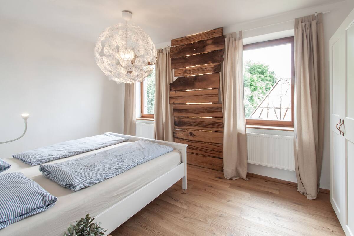 Een slaapkamer met een bed, houten paneelwand, groot raam met gordijnen en een kroonluchter.