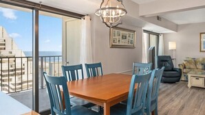 Uma sala de jantar com uma mesa de madeira e cadeiras azuis, uma poltrona de couro preto, um sofá e um abajur.
