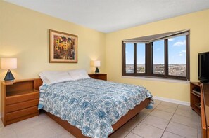 Ein Schlafzimmer mit einem Bett, zwei Nachttischen, einem Fernseher und einem großen Fenster mit Blick auf die Stadt.