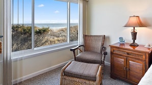 A room with a large window offering a view of the beach, a wicker chair, a small ottoman, and a wooden nightstand with a lamp. - Stunning Direct Ocean Front 3 Bedroom with Outdoor Pool! (Ocean City)