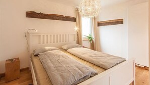 A bedroom with a white bed, a wooden headboard, a hanging light fixture, and a window with curtains. - Wohnen am Bäckerbrunnen (Würzburg)