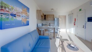 Quarto de hotel moderno com sofá azul, cozinha e uma pintura grande de uma paisagem costeira.