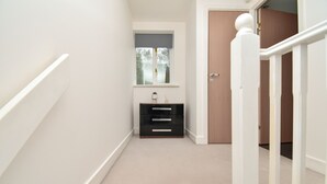 Un couloir étroit avec un tapis beige, un meuble de rangement noir doté de tiroirs, une fenêtre avec un store et une porte en bois.