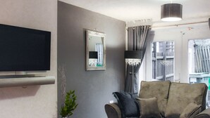 Un salon avec un canapé gris, un téléviseur à écran plat, un miroir, une lampe sur pied et une fenêtre avec des rideaux.