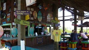 A beachside cafe with colorful stools and a variety of signs advertising free beer and surfboards.
